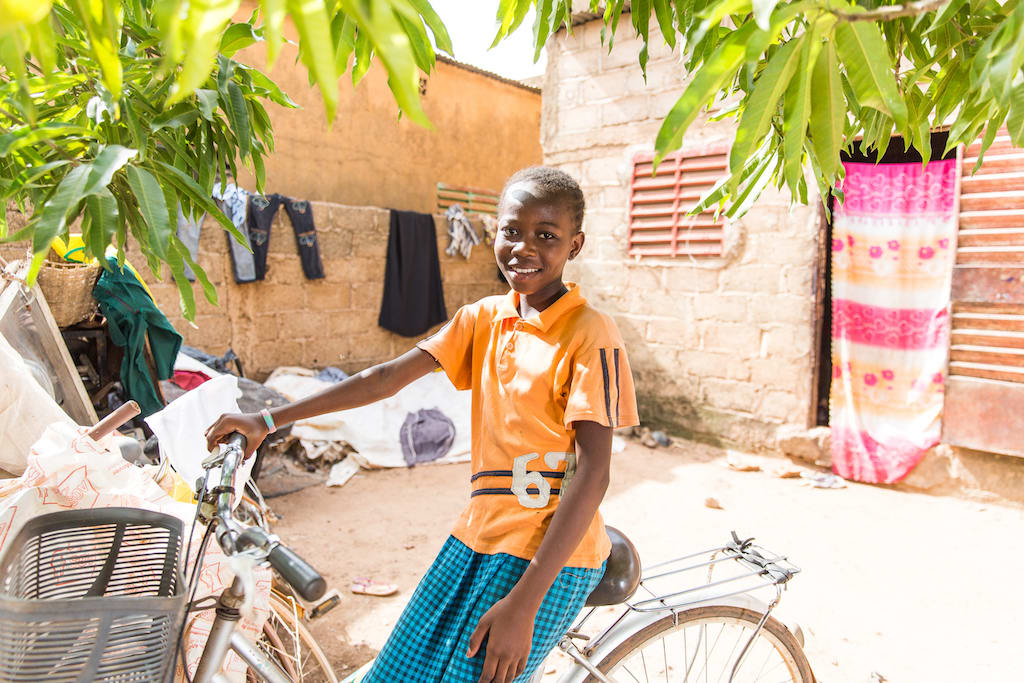 Little girl wearing an orange shirt and a green skirt smiles while sitting on her bike